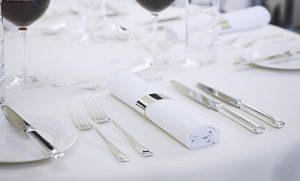 Table setting in a hotel or restaurant showing cutlery on a white linen table cloth. The knives and forks are laid either side of a napkin, wrapped nearly in a silver napkin ring.